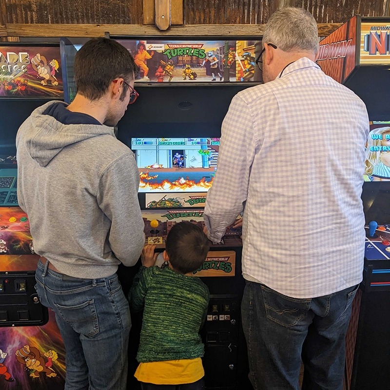 Two members of the Factory playing an arcade game with a child watching them.