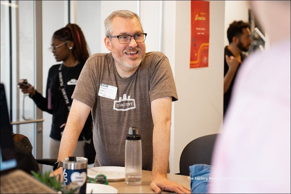 A Factory member wearing a brown shirt with a Factory brand logo, leading a conversation at a table while a community guest with braided hair and classes chats with another member.