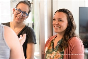 A former Factory member wearing blue framed glasses standing next to a current Factory member listen to a conversation with other Factory member.