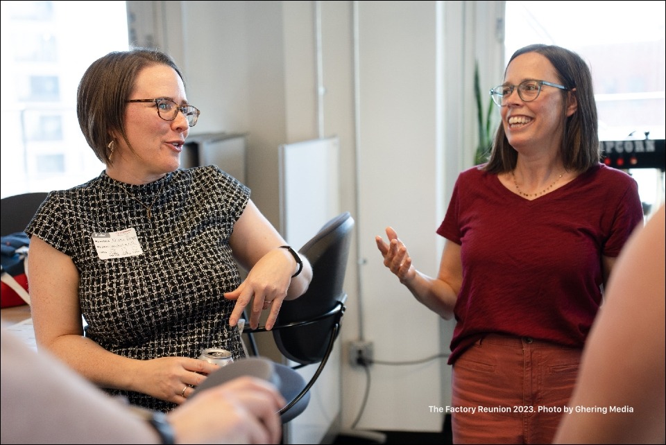 Two Factory members who are wearing glasses, one in a black and white short sleeved top and the other in a red short sleeve top, catch up on life.