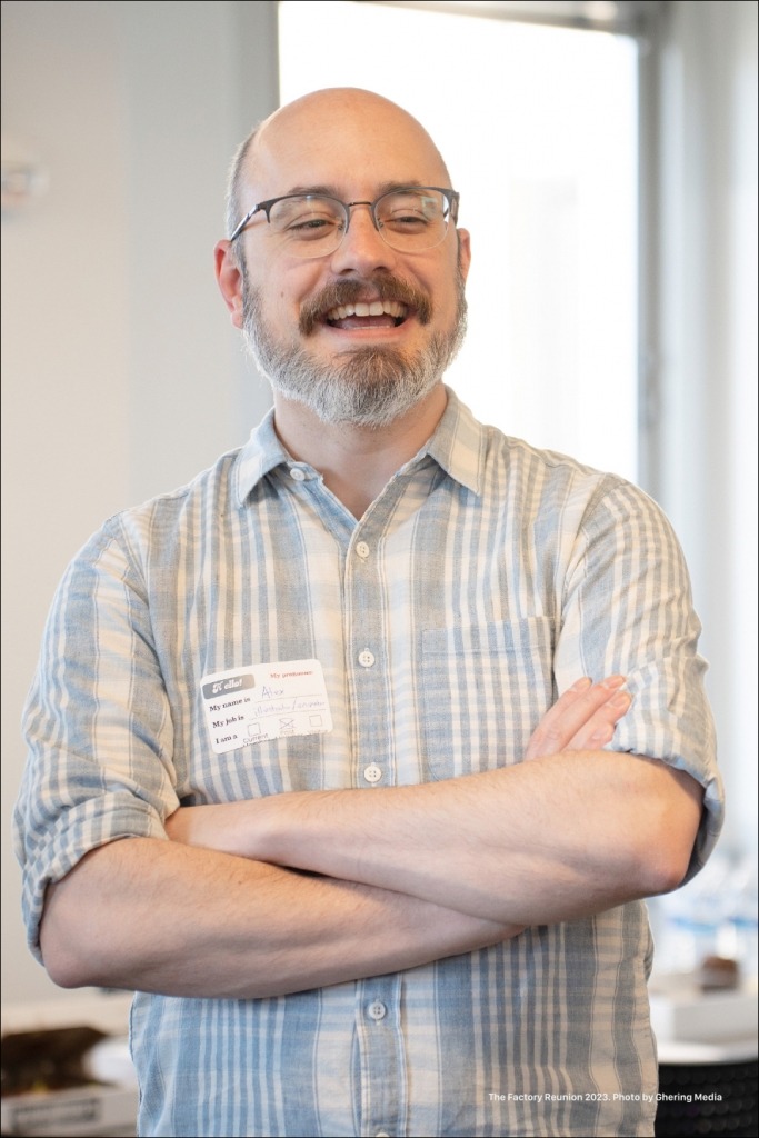 A former, bearded Factory member wearing a blue and white plaid shirt, having a great laugh telling a story.