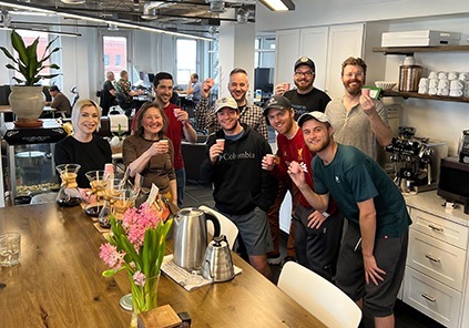 Community members gathered in the kitchen area near a large brown table top kitchen table while toasting a coffee tasting event.