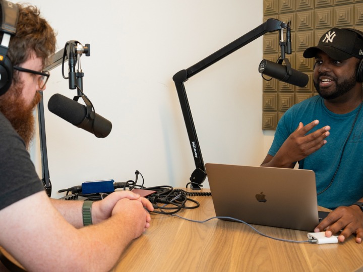 Two members of The Factory sitting at a round wooden table with microphones in front of their mouths pretending to have a discussion.