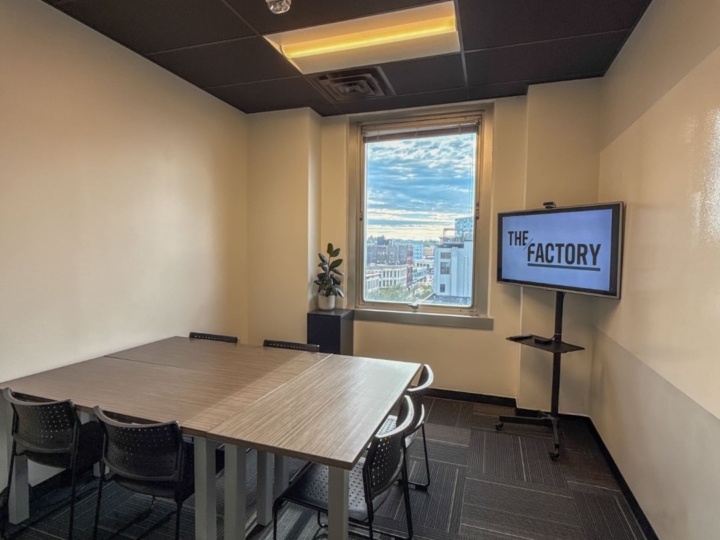 A photo of The Factory Eastown conference room with six black plastic chairs and three wooden desk placed on the north side of the room