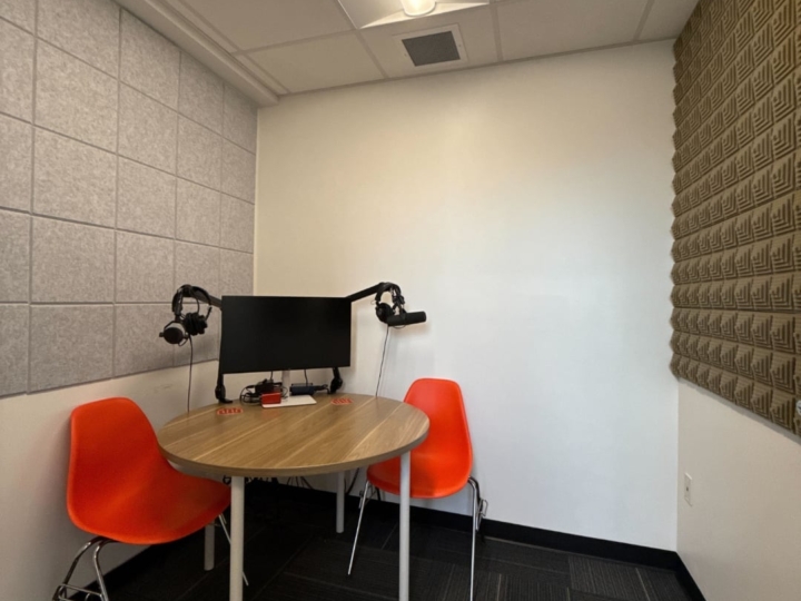 Photo of The Factory podcast booth with two orange plastic chairs and an a wooden round desk placed in the corner of the west side of the room
