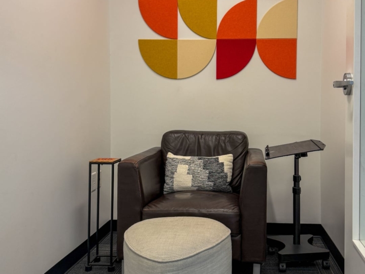 Photo of The Factory privacy and maternity booth with an large brown leather chair and light gray colored foot stool a black laptop stand and a wooden drinking cup desk