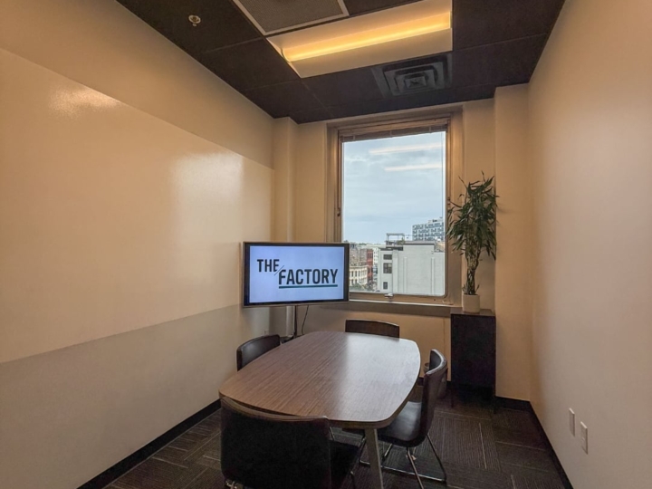 A photo of The Factory Westside conference room with four brown leather chairs and an a wooden table placed in the center of the room with a television in the northeast corner of the space.